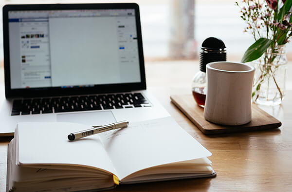 A laptop computer on a desk with an open notebook in front of it and a coffee cup to the side.